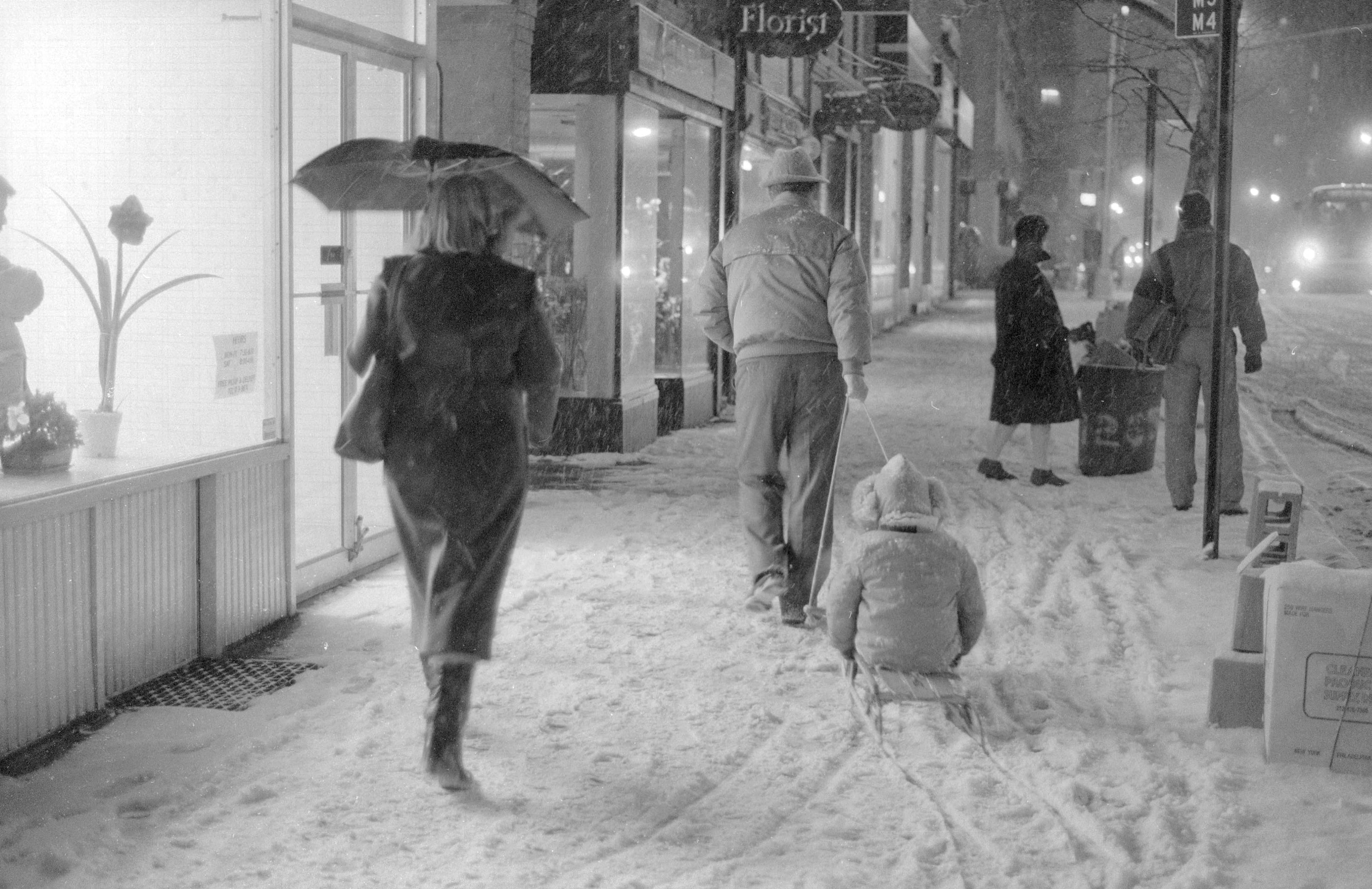 Pedestrians walk along a snowy city street at night
